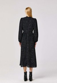 Robe noire à manches longues avec une taille froncée, présentant un motif tacheté dans des couleurs variées. Tissu doux, longueur mi-mollet, associée à des bottes à talons noires.