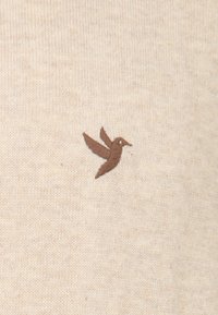 Beige gebreide stof met een zachte textuur en een klein bruin geborduurd vogelontwerp aan de linkerkant.