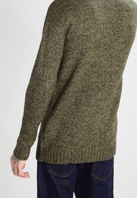 Olivgrön stickad tröja med ett texturerat mönster, ribbad fåll och ärmar, långa ärmar och en avslappnad passform, kombinerad med mörkblå jeans.