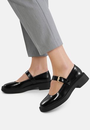 Czarne lakierowane buty typu Mary Jane z grubą podeszwą, paskiem wokół kostki i złotą klamrą, w połączeniu z szarymi, skróconymi spodniami.