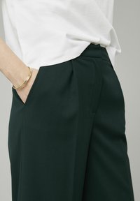 Pantalon vert foncé en tissu lisse avec des poches latérales et des plis marqués. Un haut blanc partiellement visible, avec un bracelet en or au poignet.