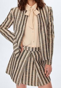 Femme portant un chemisier beige avec un nœud papillon, accompagné d'une veste et d'une jupe rayées assorties en tissu tweed noir, beige et blanc.