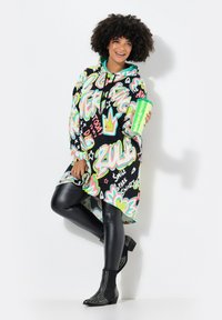 Sweatshirt surdimensionné multicolore avec des impressions graphiques, leggings noirs et bottes à talons cloutées noires. Tient une tasse verte scintillante.