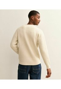 Cremefarbener gestrickter Pullover mit strukturiertem Gewebe, Rundhalsausschnitt und gerippten Bündchen. Trägt ihn mit dunkelblauen Jeans, was eine lässige Rückansicht zeigt.