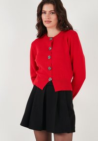 Roter Cardigan aus Strickstoff, mit tailliertem Design, rundem Ausschnitt und sechs dekorativen silbernen, rosenförmigen Knöpfen.
