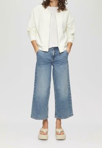 Veste bomber blanche texturée à fermeture éclair, t-shirt clair, jeans en denim bleu à jambes larges, et sandales compensées beiges. Tenue simple et décontractée.