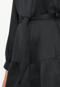Vestido negro con una tela texturizada, que presenta mangas largas, un cinturón atado en la cintura y un diseño de falda en capas. Rayas verticales visibles.