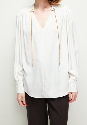 Femme portant un chemisier blanc à manches longues avec un col en V et des détails en chaîne dorée, associée à un pantalon sombre, debout devant un fond uni.