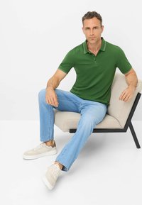 Polo verde con accenti bianchi, jeans azzurri chiari e sneaker beige. Il modello è seduto su una poltrona chiara dal design moderno.