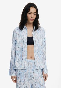 Chemise bleu clair à boutons avec des motifs floraux et des manches longues, fabriquée en tissu lisse, associée à un haut sombre et un pantalon assorti.