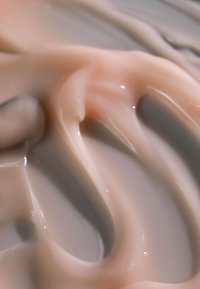 Cremige, glatte Konsistenz einer hellpfirsichfarbenen und grauen Substanz mit subtilen Wirbelmustern und Reflexionen, die auf ein kosmetisches oder Hautpflegeprodukt hinweisen.