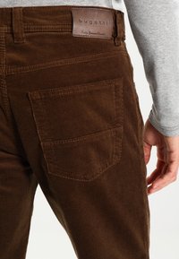 Bruna kordjeans med en texturerad yta, med en bakficka och en lädermärkesetikett märkta "bugatti" vid midjan.