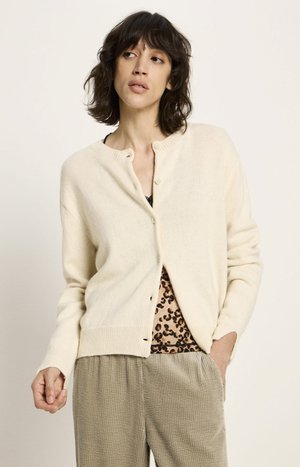Woman wearing a cream cardigan over a leopard print top and beige textured pants, looking off to the side with one hand in her pocket.