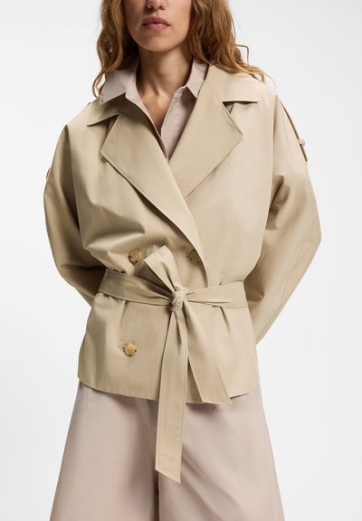 Femme portant un trench-coat beige ceinturé à larges revers sur une tenue de couleur claire, devant un fond blanc uni.