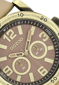 Montre ronde marron avec des accents dorés, grands chiffres et deux sous-cadrans. Elle possède un bracelet texturé et des aiguilles des heures et des minutes uniques.