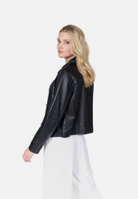 Černá kožená kráčenka s plochým límcem, výraznými švy a dlouhými rukávy, nošená přes bílé kalhoty.