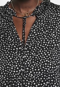 Blouse noire à fleurs avec de petites fleurs blanches, col froncé et fermeture à nouer. Tissu texturé avec une coupe légère.