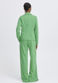 Vrouw met haar in een knot, met gouden oorbellen en een felgroen, op maat gemaakt pak, die met haar rug naar voren staat tegen een effen witte achtergrond.