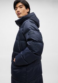 Kék pufi dzseki kapucnival, steppelt mintázattal, oldalsó zsebekkel és logóval ellátott hüvelybugyival. Simább, fényes szövettextúra.