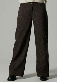 Pantalons larges marron foncé avec de légères rayures zèbre, portés avec des chaussures noires et un pull beige dont l'ourlet est visible.