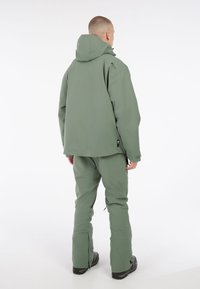 Set van maslinasto zelenim vanjskim odijelom koji se sastoji od jakne s kapuljačom i odgovarajućih hlača. Oba komada imaju glatku teksturu i funkcionalne zip ventile na gležnjevima.