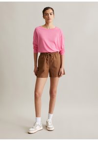 Junge Frau trägt ein pinkfarbenes Langarmshirt, braune Shorts mit Kordelzug, weiße Socken und beige Turnschuhe und steht vor einem schlichten Hintergrund.