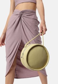 Rund gul väska med en texturerad yta, hålls av ett band. Modellen bär en texturerad lavendelfärgad kjol med en sprund framtill och knytning.