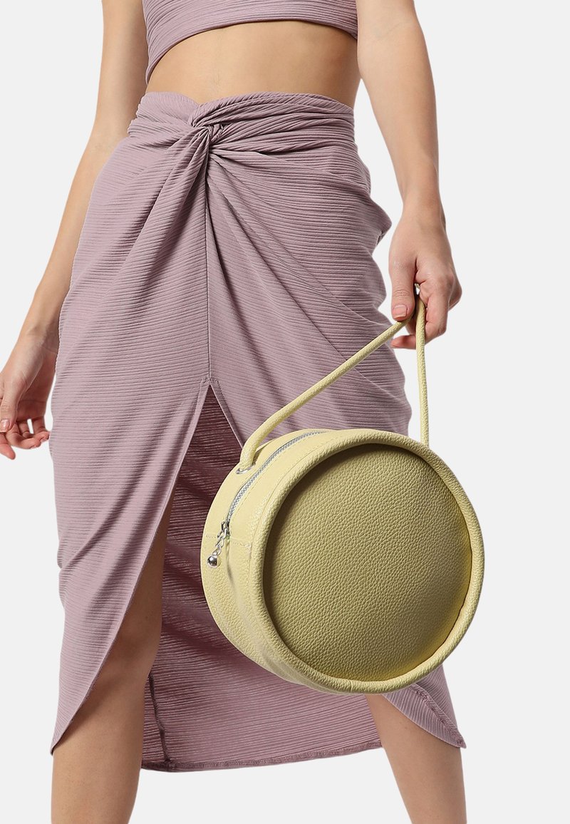 Rund gul väska med en texturerad yta, hålls av ett band. Modellen bär en texturerad lavendelfärgad kjol med en sprund framtill och knytning.