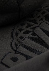 Crni pamučni materijal s teksturiranim, tamnim izvezenim dizajnom koji sadrži geometrijske uzorke i slova, stvarajući suptilan kontrast.