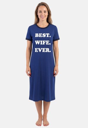 Smilende kvinde står barfodet iført en blå midi-kjole med korte ærmer og hvid tekst foran, der lyder "BEDSTE. KONE. NOGENSINDE."