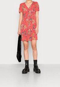 Robe courte rouge à motif floral, décolleté en V et manches bouffantes. Associée à des bottes à plateforme noires jusqu'au genou et à un sac en fausse fourrure noir.