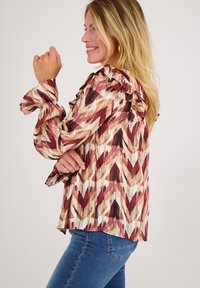 Blouse met lange mouwen en een meerkleurig zigzagpatroon in rood, crème en bruin. Bevat ruches aan de schouders en manchet.
