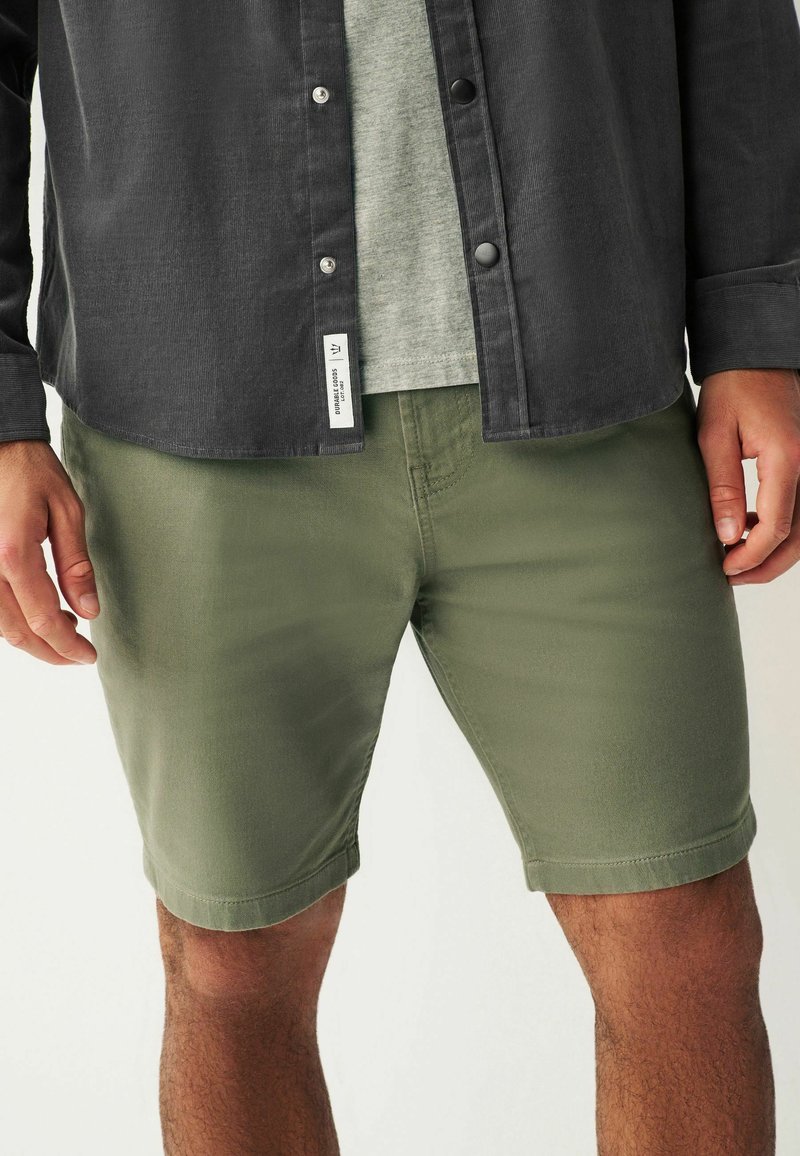 Shorts vert olive en coton, avec une coupe slim et un ourlet roulé. Associés à un t-shirt gris et une chemise à boutons foncée.