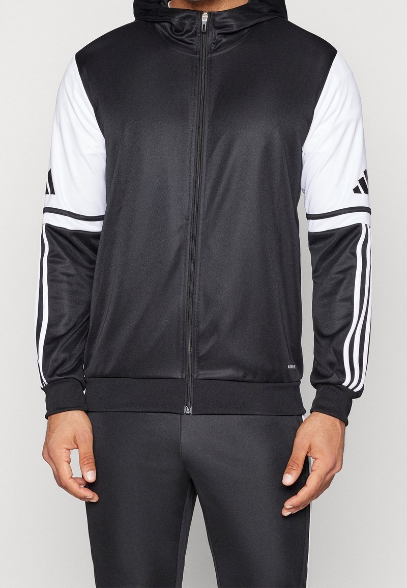 Homme portant une veste de sport noire et blanche zippée avec capuche et un pantalon de sport noir assorti.