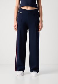 Frau in navyblauen, weiten Hosen mit roten und weißen Seitenstreifen, weißen Sneakers und einem navyblauen Croptop, die vor einem schlichten Hintergrund steht.