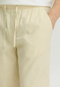 Beige, lette bukser med elastisk midjebånd og snøring. Side lommer er synlige. Stoffet har en glatt, myk tekstur.