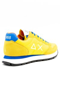 Žlutá sportovní bota s velurovou a textilní svrškem, modrými akcenty, bílým logem a černou gumovou podešví. Disponuje polstrovaným límcem a kontrastními tkaničkami.