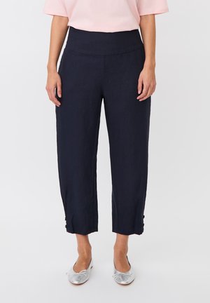 Frau trägt marineblaue Cropped-Hosen mit Knopfdetails am Saum, hellrosa Kurzarmtop und silberne Ballerinas, steht vor weißem Hintergrund.