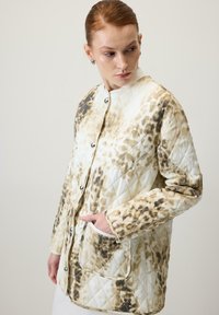 Nő egy bézs és barna állatmintás, steppelt, gombos kabátban, egyik kezével a zsebben, oldalt nézve egy sima háttér előtt.