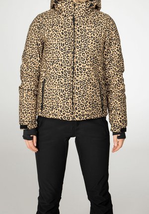 Persoon die een beige en zwarte pufferjas met luipaardprint en capuchon draagt, en zwarte broek, staand tegen een effen lichte achtergrond.