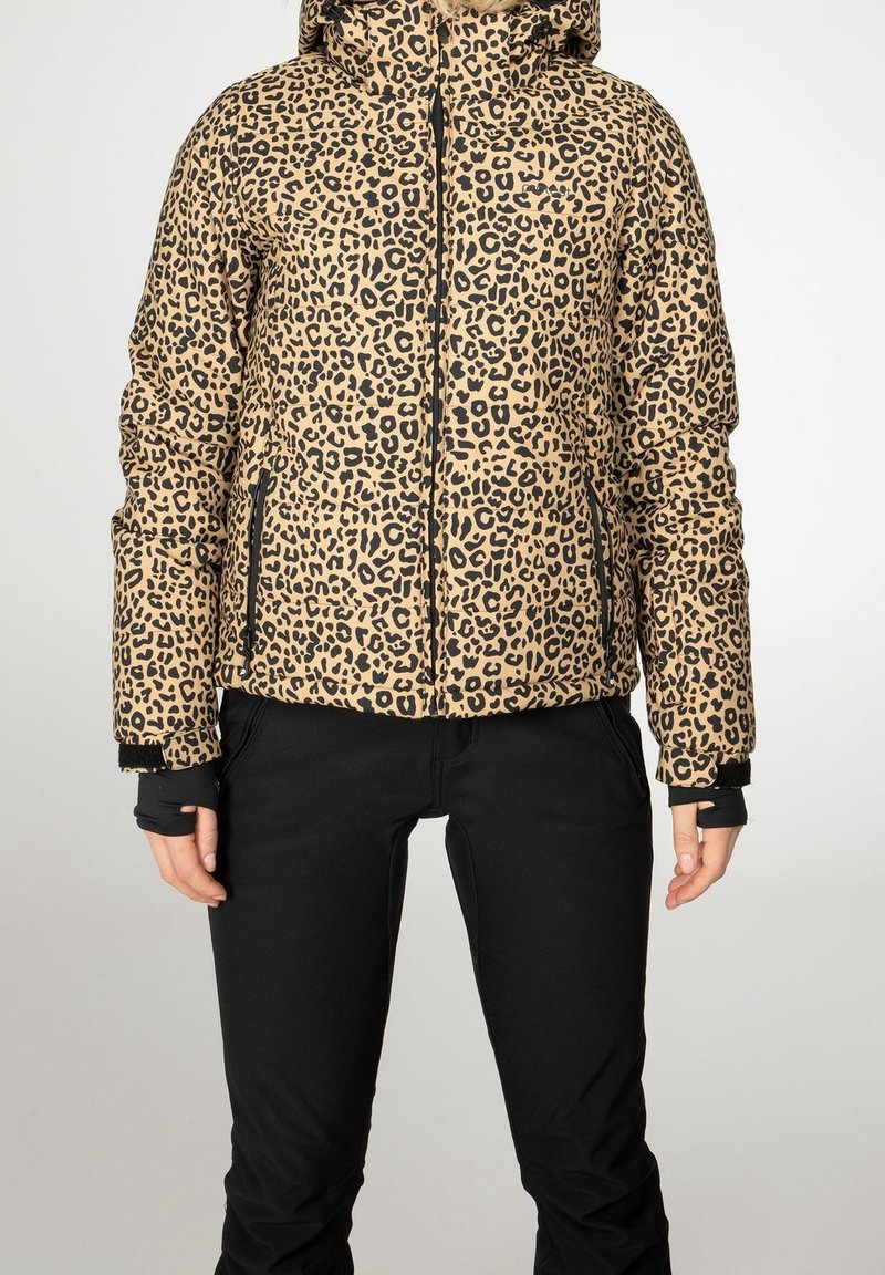 Personne portant une doudoune beige et noire à imprimé léopard avec capuche et pantalon noir, debout devant un fond clair uni.