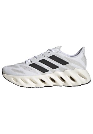 Zapatilla deportiva blanca con logotipo de tres franjas negras, parte superior de malla con textura, cuello acolchado y diseño distintivo de la suela en forma de ondas.
