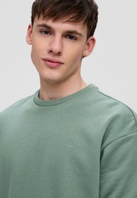 Felpa verde chiaro realizzata in materiale morbido, con collo rotondo e discreto logo QS sul petto. Tessuto liscio con vestibilità rilassata.