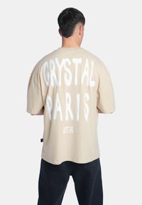 Béžové oversized tričko s v ýrazným bílým textem "CRYSTAL PARIS" na zádech, s dalším textem "DEPT. 75" pod ním. Krátké rukávy.