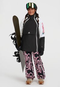 Zwart-witte waterdichte jas met ritszak, gecombineerd met kleurrijke geprinte broeken. Houdt een snowboard vast en draagt skibril.