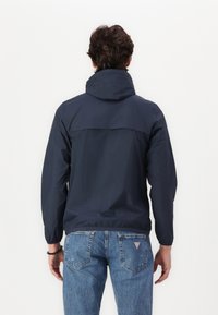 Navy windbreaker jacket with a high collar, elastic cuffs, and a relaxed fit. Paired with faded blue jeans, featuring a triangular logo on the pocket.