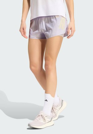 Kvinde iført lyse lilla træningsshorts, hvide Adidas-sokker og beige løbesko, stående mod en ensfarvet lys baggrund.