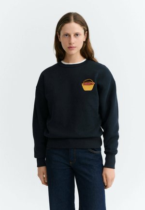 Femme aux longs cheveux bruns porte un sweat-shirt noir avec un petit panier pique-nique brodé sur la poitrine et un jean bleu foncé.