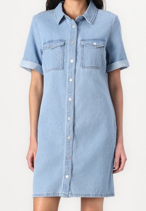 Jasnoniebieska sukienka z krótkim rękawem z denimu, z przodu zapinana na guziki, z dwoma kieszeniami na piersiach z klapkami, noszona przez osobę stojącą na jednolitym tle.