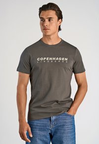 Joven con cabello oscuro que lleva una camiseta gris "Copenhague Lindbergh" y jeans azules, con una mano en el bolsillo, mirando hacia la derecha.